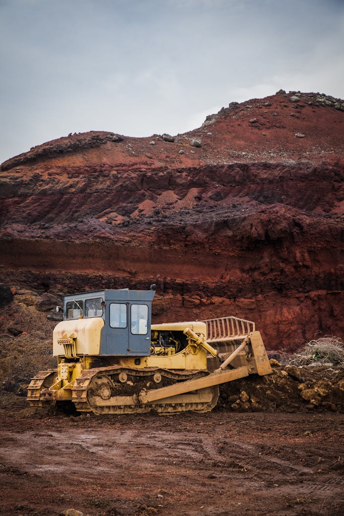 A yellow bulldozer excavating in a rugged red earth quarry under a cloudy sky.